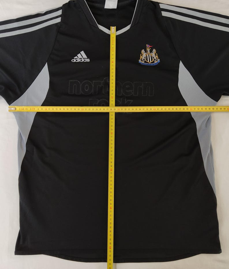 May include: Black and grey Adidas Newcastle United football shirt. The shirt displays the Adidas logo, the Newcastle United crest, and the text "northern rock". It has grey stripes on the shoulders.