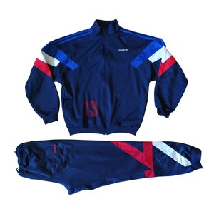 Può includere: Un completo tuta blu navy con dettagli rossi e bianchi. La giacca ha una chiusura con cerniera e un logo Adidas bianco sul petto. I pantaloni hanno una cintura elastica con coulisse e una striscia rossa e bianca lungo il lato. Il numero "13" è ricamato sulla giacca.