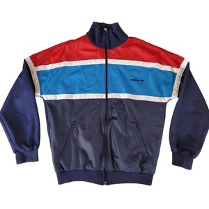 Op de afbeelding: Een vintage Adidas trainingsjack in marineblauw, rood en lichtblauw. De jas heeft een volledige ritssluiting, een hoge kraag en het Adidas-logo op de borst. De mouwen en de onderkant zijn marineblauw, met rode en lichtblauwe strepen.