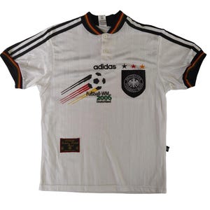 Op de afbeelding: Wit Adidas voetbalshirt met zwarte strepen op de schouders en manchetten. Voorzien van het Duitse nationale embleem, Adidas logo en tekst "Fußball-WM 2006 Deutschland". Het shirt heeft een kraag met knopen en een kleine patch linksonder.