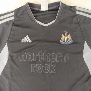 May include: Black and grey Adidas football shirt featuring the Newcastle United crest and the 'northern rock' logo. The shirt has grey stripes on the shoulders and a V-neck collar.