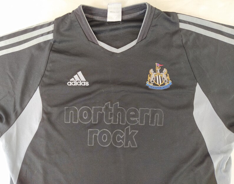 May include: Black and grey Adidas football shirt featuring the Newcastle United crest and the 'northern rock' logo. The shirt has grey stripes on the shoulders and a V-neck collar.