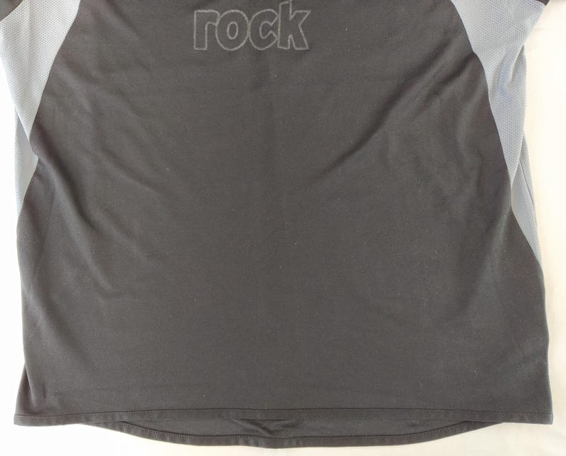 May include: Black athletic shirt with grey mesh side panels. The word "rock" is embroidered across the upper back in a darker shade of grey. The shirt has a crew neck and short sleeves.