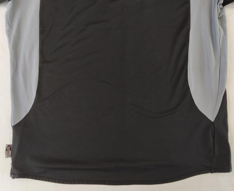May include: Black athletic top with grey mesh side panels. The shirt features a curved hem and a small tag on the lower left. The fabric looks like moisture-wicking material, suitable for sports and exercise activities.