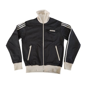 Puede incluir: Chaqueta deportiva negra con rayas blancas en las mangas y cuello y puños crema. La chaqueta tiene cremallera completa, dos bolsillos delanteros y el logo de Adidas en el pecho.