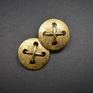 May include: Two round, gold-colored buttons with a textured surface and a cross-shaped design. Each button has four holes and a raised cross detail. The buttons are set against a dark gray background.