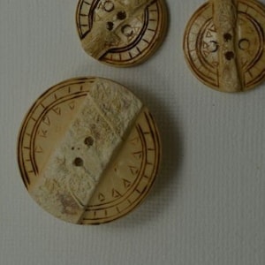 May include: Three round, wooden buttons with a decorative design. The buttons are light brown and have a geometric pattern carved into them.