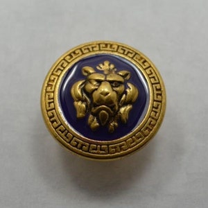 May include: A gold-colored button with a blue enamel center featuring a lion's head design. The button is surrounded by a gold-colored border with a Greek key pattern.