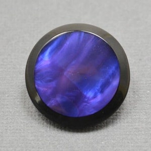 May include: A round, black button with a purple and blue marbled design. The button has a raised edge around the outside.
