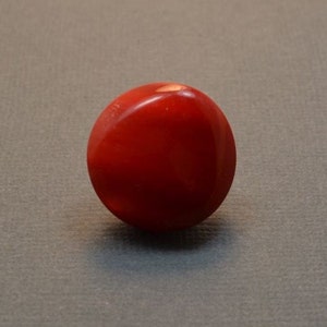 May include: A smooth, round, red gemstone. The gemstone has a slight sheen and is polished.