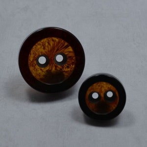 May include: Two brown plastic buttons with black rims and white centers. The larger button has a swirling pattern on the surface.