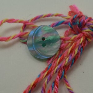 May include: A blue button with a pearly finish and two holes, tied with a multi-colored yarn in pink, yellow, blue, and purple.