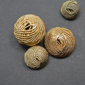 May include: Four gold-colored buttons with a woven design. The buttons are round and have a raised center.