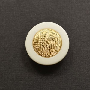 May include: A white button with a gold, textured center. The button has a raised edge around the center.
