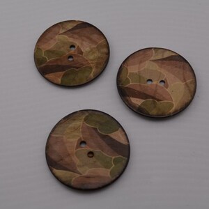 May include: Three large, round wooden buttons with a camouflage pattern in shades of brown, green, and tan. Each button has two small holes for sewing. The buttons are arranged on a light gray surface.