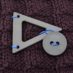 May include: A black triangular button with blue thread and a black round button with a blue thread loop. The buttons are on a brown knitted fabric.