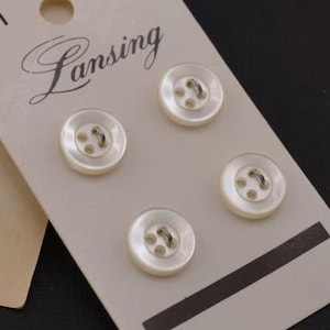 May include: Four white plastic buttons with four holes each and a silver shank. The buttons are on a white card with the word "Lansing" printed on it.