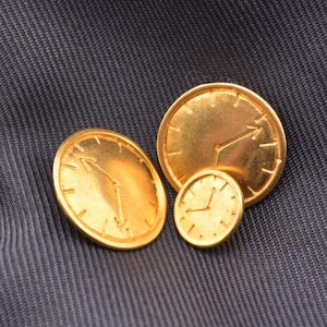 May include: Three gold-tone metal clock face buttons with black hands. The largest button is about 2 inches in diameter, the middle button is about 1.5 inches in diameter, and the smallest button is about 1 inch in diameter.