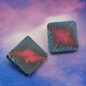 May include: Two square-shaped earrings with a black and blue swirl pattern and a pink center.