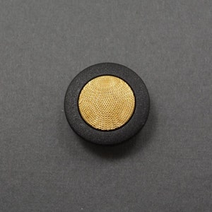 May include: A black button with a gold, textured center. The button has a raised edge around the center.