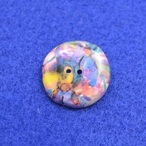 May include: A colorful, abstract button with two holes. The button is round and has a white background with splashes of pink, yellow, blue, and purple paint.