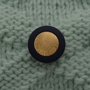 May include: A black button with a gold center, featuring a textured design. The button is on a light green knitted fabric.