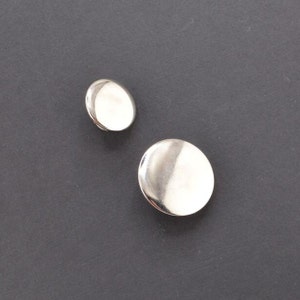 May include: Two silver-toned, round, flat buttons. The larger button is slightly larger than the smaller button.