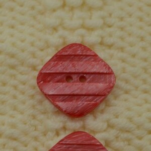 May include: Two red plastic buttons with a striped pattern and two holes. The buttons are on a white knitted surface.