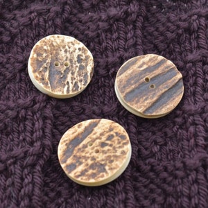 May include: Three round, brown and white patterned buttons with two holes each. The buttons are on a purple knitted fabric.