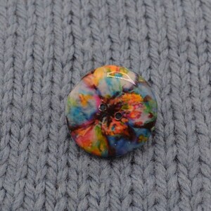 May include: A round, colorful button with a tie-dye pattern in shades of blue, pink, yellow, and orange. The button has two black holes for sewing.