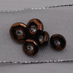 May include: Five brown buttons with a shiny finish and clear rhinestones in the center. The buttons are on a silver and white fabric background.