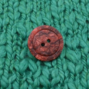 May include: A round, reddish-brown button with two holes, set against a textured, teal green knitted background. The button has a marbled appearance, with darker brown swirls within the reddish-brown material. The button is a sewing or craft supply.