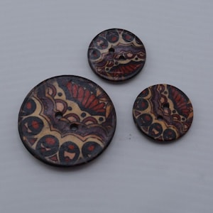 May include: Three round wooden buttons with a colorful mandala design. The buttons are brown, beige, red, and purple. The largest button is about 2 inches in diameter.