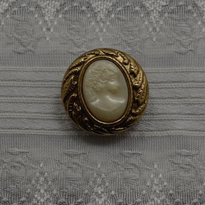 May include: A gold-toned button with an oval cameo of a woman's face. The cameo is surrounded by a decorative floral design.