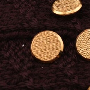 May include: Close-up of four gold-colored round buttons with a textured surface. The buttons are on a dark brown fabric.
