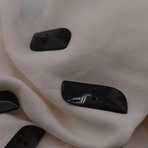 May include: Four black, shiny, plastic buttons with two holes each. The buttons are shaped like a slightly curved rectangle.