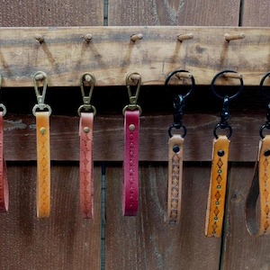 May include: Seven leather keychains hanging in a row. Four feature brass-toned swivel clasps and embossed geometric patterns in red, tan, and pink. Three feature black metal rings and clasps with tan leather straps and intricate stamped designs.
