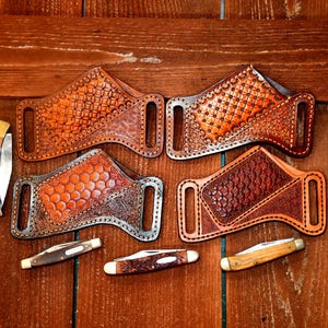 May include: Four brown leather knife sheaths with distinct geometric embossed patterns and stitched edges, each featuring two belt loops. Four folding pocket knives with various handle materials including wood and black plastic are arranged around them.