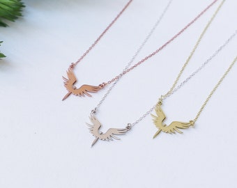 May include: Four delicate necklaces with a winged pendant design in rose gold, silver, and gold tones. The necklaces are displayed on a white surface with green foliage in the upper left corner. The pendants have a stylized bird or angel wing shape.