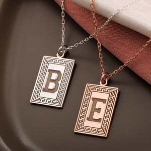 May include: Two rectangular pendant necklaces featuring the letters B and E. The silver-coloured pendant displays the letter B, while the rose gold-coloured pendant displays the letter E. Both pendants feature a Greek key border pattern and thin chains.