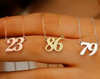 May include: Three gold necklaces with different numbers, 23, 86, and 79, hanging on a hand. The necklaces are made of different metals, with the 23 necklace being rose gold, the 86 necklace being yellow gold, and the 79 necklace being silver.