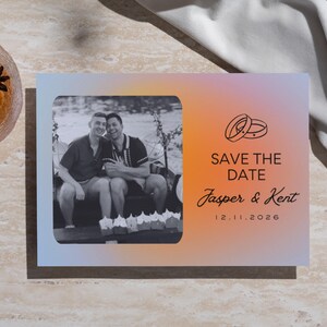 Peut inclure: Carte de faire-part avec un fond dégradé orange, rose et bleu. La carte présente une photo en noir et blanc de deux hommes, avec le texte "SAVE THE DATE" et les noms "Jasper & Kent" ainsi que la date 12.11.2026. Un verre de liquide ambré avec de l'anis étoilé est dans le coin supérieur gauche.