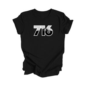 Op de afbeelding: Een zwart T-shirt met korte mouwen en de witte tekst 716 op de borst. De cijfers zijn gevuld met een zwarte silhouet van een stadsskyline, wat een grafisch ontwerp creëert dat contrasteert met de zwarte stof van het kledingstuk.