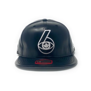 May include: Black leather baseball cap with a white embroidered logo featuring a stylised number "6" with an eye and a Canadian maple leaf inside. The cap has a black adjustable strap with a red "Snapback" label.