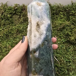 May include: A polished, green and white moss agate crystal tower. The crystal has a pointed top and a hollowed-out section with small crystal formations. The stone is held against a background of green moss.