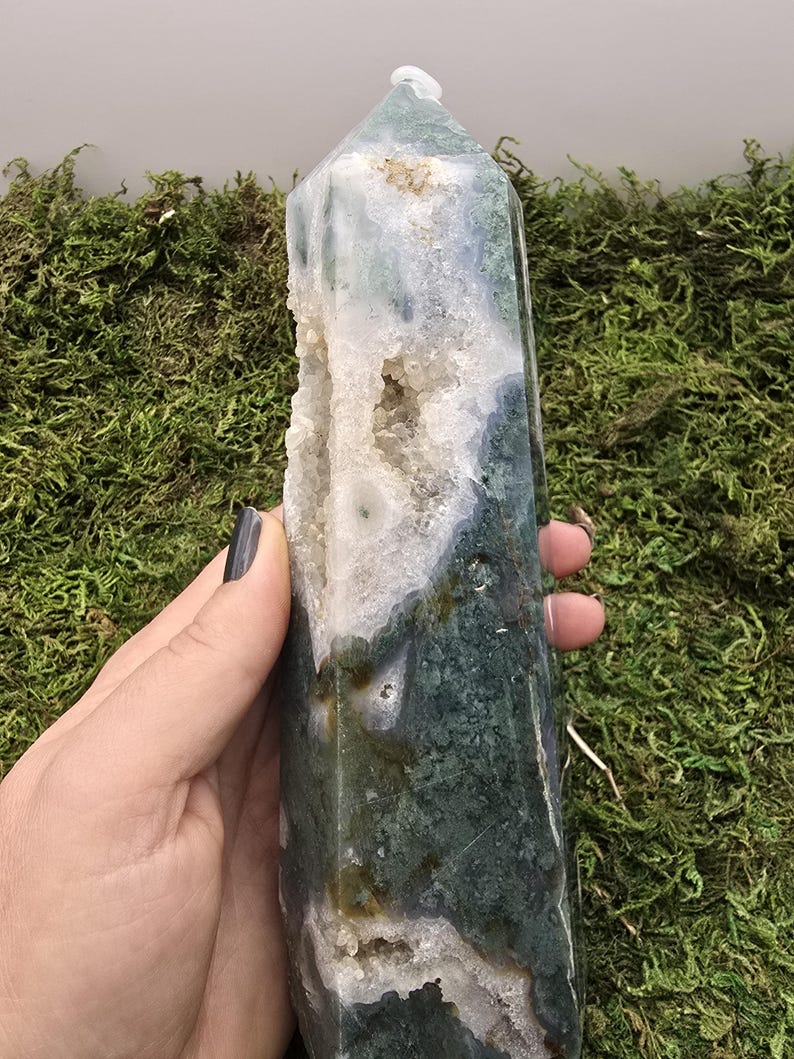 May include: A polished, green and white moss agate crystal tower. The crystal has a pointed top and a hollowed-out section with small crystal formations. The stone is held against a background of green moss.
