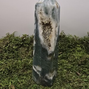 May include: A tall, polished crystal tower with a geode cavity. The crystal features a mix of green, white, and gray hues, with a central opening revealing a cluster of small, sparkling crystals. The tower is displayed on a bed of green moss.