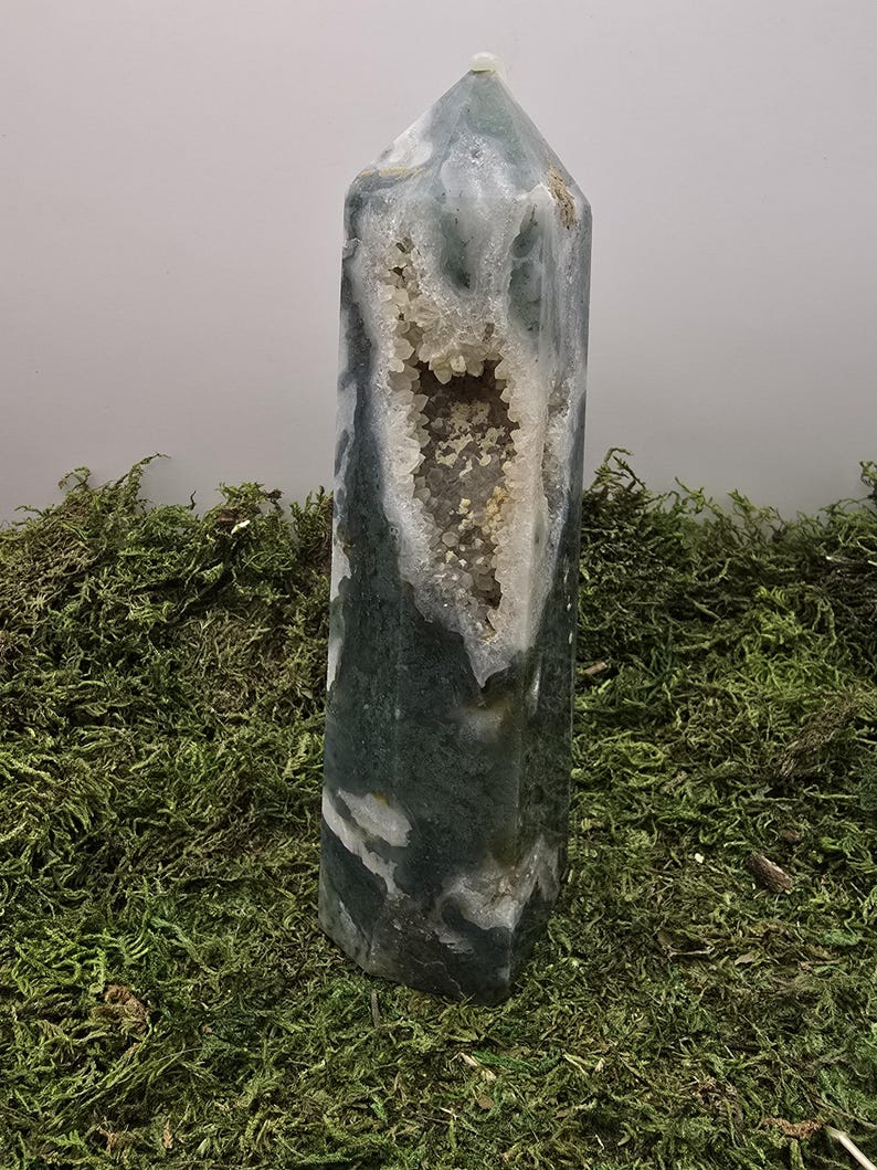 May include: A tall, polished crystal tower with a geode cavity. The crystal features a mix of green, white, and gray hues, with a central opening revealing a cluster of small, sparkling crystals. The tower is displayed on a bed of green moss.