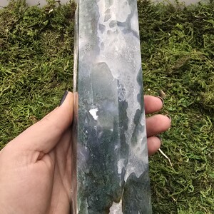 May include: A tall, polished crystal obelisk with a rounded top. The stone features a mix of white, green, and gray patterns. The obelisk is held against a backdrop of green moss.