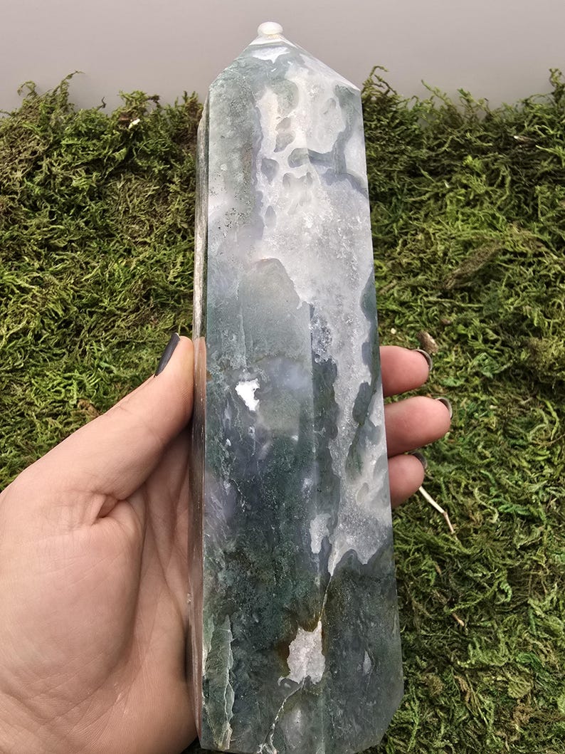 May include: A tall, polished crystal obelisk with a rounded top. The stone features a mix of white, green, and gray patterns. The obelisk is held against a backdrop of green moss.